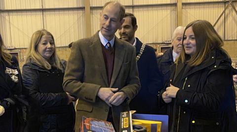 The Duke of Edinburgh stands amongst five people in front of trolleys of children's' toys. He is wearing a green blazer, burgundy sweater and white shirt. Those around him are wearing dark coloured coats.