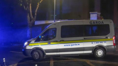 A white Garda van with a yellow stripe along its side pulls out onto a main road on a dark night.