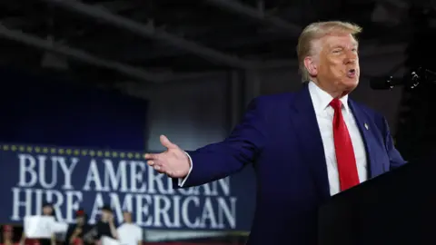 Donald Trump speaking at a microphone in front of a sign reading 'buy American, hire American'