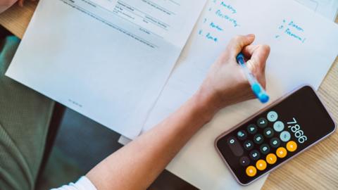 A person writing notes about expenses on a piece of paper next to a calculator