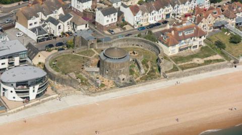 The circular Sandgate Castle, surrounded by a stone wall, located on a large sandy beach. The castle is seen from above and surrounded by other, more modern buildings.