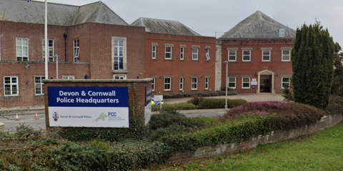 The Devon and Cornwall Police Headquarters building is a series of adjoining red brick, two-storey buildings. There are many windows and the buildings have grey slate roofs. There is a sign at the front which reads "Devon and Cornwall Police Headquarters" and bears the force's logo which is a map of Devon and Cornwall. There is a planted flowerbed in the foreground and a lawn.