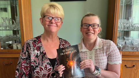 Zoë Castle and her mum Di sitting together in front of a cabinet filled with glass bowls and cups. Zoë has her hair tied back and is wearing glasses. She is holding up a book which is black with an orange heart in the centre. Di has short blonde hair and a floral patterned top. 
