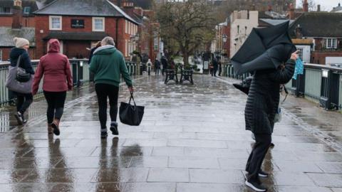 Four people walking over a bridge in the rain. On the right hand side, someone is struggling with an umbrella that has turned inside out in the wind.