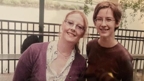 A mum stands with her grown-up daughter as both smile into the camera. The mother has strawberry blonde hair and is wearing a purple cardigan, while her daughter wears a brown top. Both are wearing glasses.