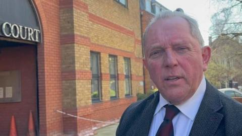 Derek Bennett: a grey haired man wearing a grey jacket and red tie outside a red-and-brown brick building. The word "court" is written above the entrance to the building in capital letters.