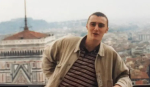 A young man with close cropped hair and beige jacket stands against a city backdrop.