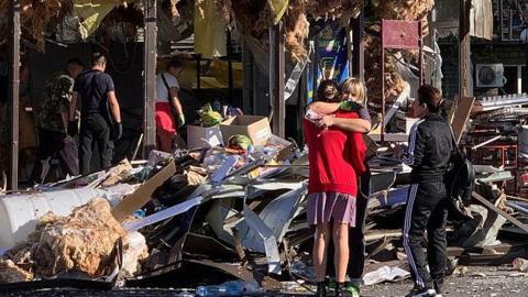 Two women hug on street filled with debris, damaged building behind them after Russian strike, a woman stands next to them and three people walk through the rubble behind them.