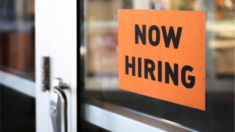 Now hiring sign on the glass front door of a business. The sign is orange with black writing.