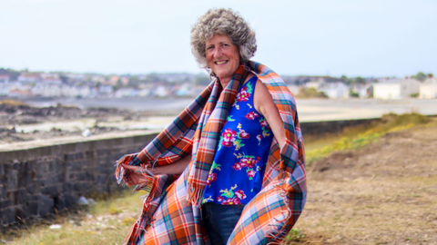 A woman wearing a tartan blanket around her arms and shoulders standing on grassland next to the beach. Rows of homes can be seen on the curved island in the distance.