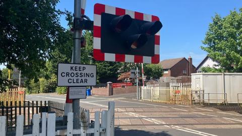 General view of the crossing with rail lines going across the road and to the left three lights in a black box surrounded by white and red stripes to the side of a sign that says 'keep crossing clear'