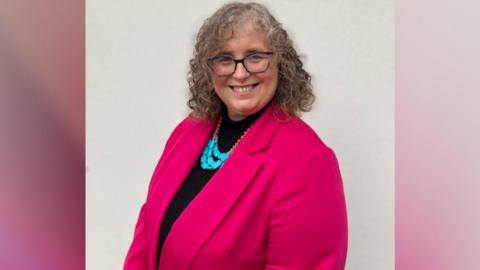 Anna Smith, who has shoulder-length brown and grey hair and is wearing glasses, smiles at the camera. She is wearing a a pink jacket and a top. 