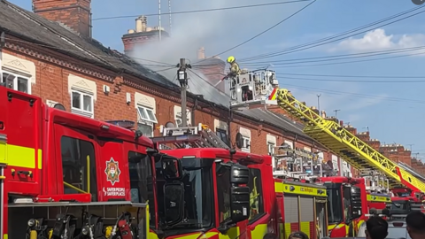 Numerous fire engines in a narrow terrace street. A firefighter is standing on an aerial ladder platform using a water hose to tackle a fire in a roof.