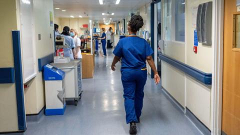 General view of a hospital ward - a medical professional walks down the middle- with other staff in masks on the left 