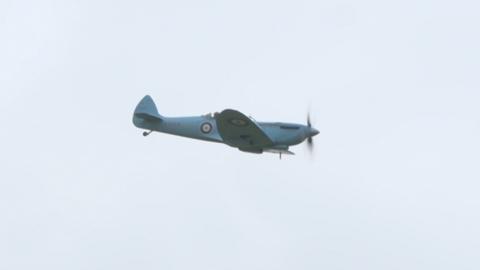 A Spitfire painted in a pale blue colour with a circular logo on the side flies through the air.
