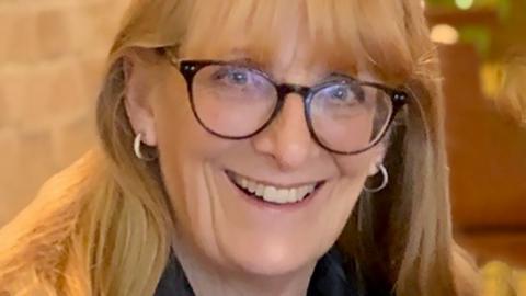 Margaret Parker is a blonde haired woman and is wearing plastic-rimmed glasses. She is smiling.

