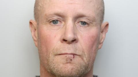 Darren Morris looks into the camera for his custody image. He has a small cut to his lip. He has short facial hair, which is light grey and ginger-y, and short hair of the same colour.