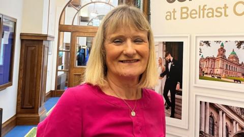 A woman with mid length blonde hair smiles into the camera. She is wearing a pink top and a gold necklace with a pendant. She is standing in front of a board displaying marriages at Belfast City Hall.