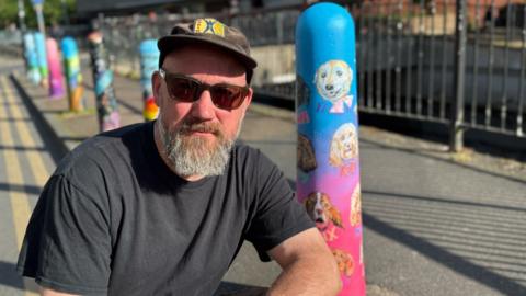 A man crouching next to a bollard painted blue and pink with portraits of dogs.