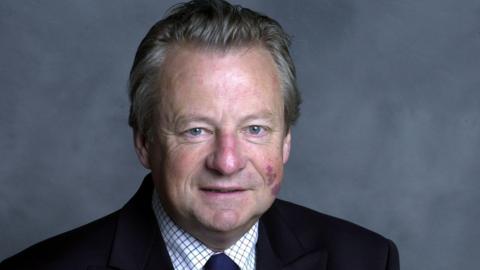 Dafydd Elis-Thomas who has grey hair and a birthmark on his cheek. He is wearing a black suit and is smiling and looking at the camera.