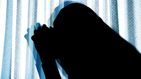 Shows a silhouette of a woman kneeling with her hands clasped in front of her head.