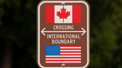A close-up of a road sign that reads "Crossing International Boundary". Above the words, there is a white and red Canadian flag. Below the words, there is a red, white and blue American flag.