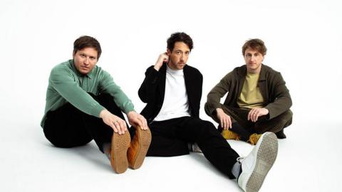 The Wombats sitting on the floor (left to right) Tord Overland Knudsen wearing a green shirt and black trousers holding the toes of his trainers, Matthew Murphy scratching behind his ear wearing a white t-shirt and black jacket and black trousers and white trainers and Dan Haggis sitting cross-legged wearing mustard-coloured t-shirt and dark green shirt and trousers and mustard-coloured trainers