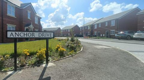 A sign reads Anchor Drive as the road leads round into a residential area - with houses on both sides of the road and cars in the drive