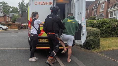 Two police officers, one in a black uniform and helmet and one in a white T-shirt and black vest, push a man in a green jumper into the back of a police van