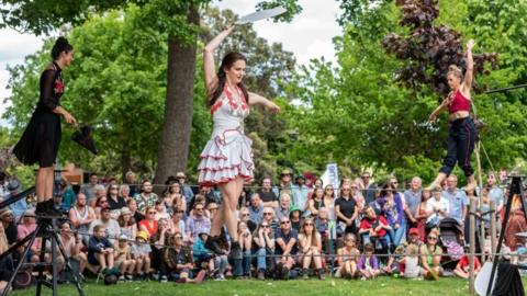 A troupe of three female wire walkers perform in front of a crowd in a park. The woman closest to the camera is holding a machete knife while balancing on the wire. 