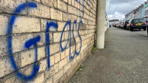 A brick wall with the word "grass" in dark blue graffiti paint. There is a pavement in front of it with a lamppost, and cars parked along the side.