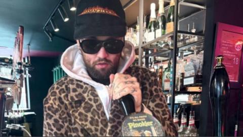 Mason Newman, who has a beard, stands behind the bar and pulls a pint. He is wearing shades and a leopard-print coat, as well as a black baseball cap. He is looking at the camera.