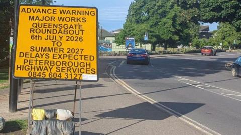 A road sign on approach to a roundabout. It has black lettering on a yellow background and says: "ADVANCE WARNING. MAJOR WORKS. QUEENSGATE ROUNDABOUT. 6TH JULY 2026 TO SUMMER 2027. DELAYS EXPECTED. PETERBOROUGH HIGHWAY SERVICES. 0845 604 3127."