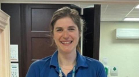 A woman wearing a blue NHS top and a lanyard is smiling at the camera. She has dark hair.
