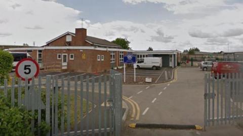 School buildings with parked vans and cars, a gate partially open leading in with a speed bump; a 5mph sign and cloudy skies in the background.