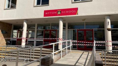 Image shows the exterior of Myton School, and includes the front entrance and sign saying 'Myton School,' along with its logo which is a shield. The writing is on a red background, and the sign is stuck to a white wall.