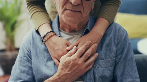 Shot of bottom half of older man's face and upper body. There are arms from behind comfoting him