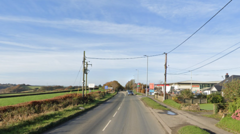 A road with two carriageways and some telegraph poles and cables overhead and some buildings on the right and fields on the left.