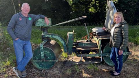 David wears a grey top with the Battisford & District club logo on it, while Julia wears a green jacket and denim jeans. They are standing at either end of a green old cricket pitch roller, in front of some trees.