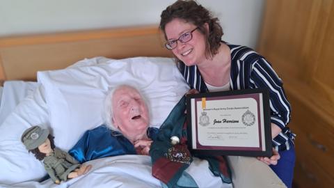 A picture of Joan lying in bed being presented a certificate. She is next to a toy and another woman who is presenting the certificate.