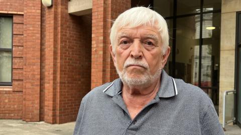 Raymond Beeson stands outside Hull Crown Court. He is wearing a grey, black and white polo shirt. He has white hair and a beard. 