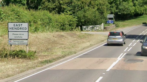 The A417 Reading Road in East Hendred. It is a wide single lane road. There is a white sign next to the street that reads 'East Hendred'.