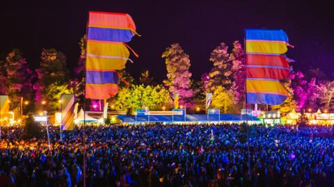 Fields of festival attendees at Kendal Calling in 2024. There are flags in the foreground and stalls in the background. Between stand thousands of people. The shot is taken in the evening and a purplish-blue glow lights up the crowd.