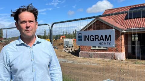A man standing in front of a building site. He is wearing a blue shirt and has brown hair. On the metal fence is a sign for his company, "Ingram Homes".