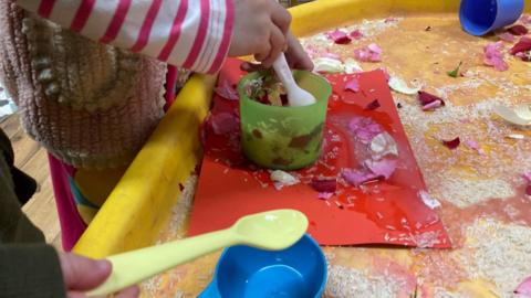 Young children's hands playing with spoons and buckets. There is rice spilled out on a yellow table.