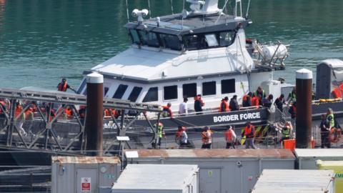 Border force boat with migrants walking on board wearing life jackets 