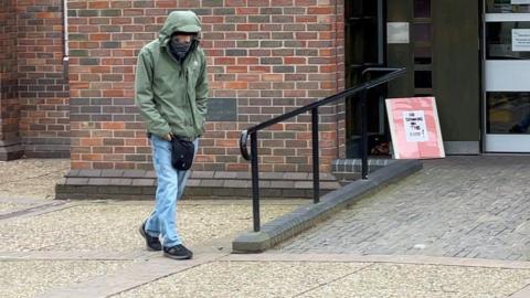 Christopher McEwen walks near the entrance of a building. He is wearing light blue jeans, with a green jacket with the hood up. He has a scarf covering the lower part of his face. He is hunched over with his hands in his pockets. 