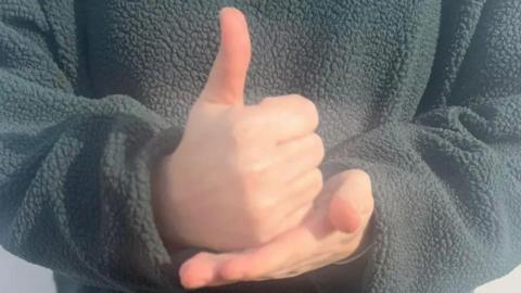 A close-up of a person signing - it shows a flat palm, with the other hand placed on top giving a thumbs up, the person is wearing a grey fleece top.