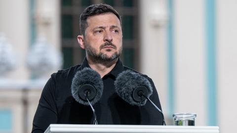 Zelenksy looks pensive as he focuses his attention to the right of frame while he stands at a podium against a light blue bokeh background, on the podium are two microphones and a glass of water.