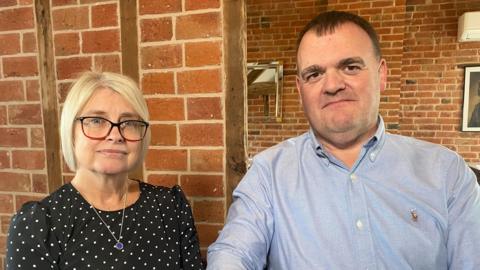 Sue and Dave sat side by side with serious expressions. Sue is wearing a black dress with white dots, and Dave is wearing a blue button-up shirt. They're sat indoors in front of a red brick wall. 
